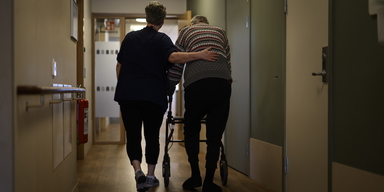 En person går med rullator med ett vårdbiträde bredvid på ett omsorgsboende för äldre. När en kommun inte kan erbjuda plats på särskilt boende inom en skälig tid kan den få betala en straffavgift.