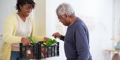 En kvinna håller fram en back med grönsaker och en äldre man tittar på dem. Du kan förbättra din hälsa så att du åldras bättre, även om du hunnit fylla 65, en växtbaserad kost är en bra start.