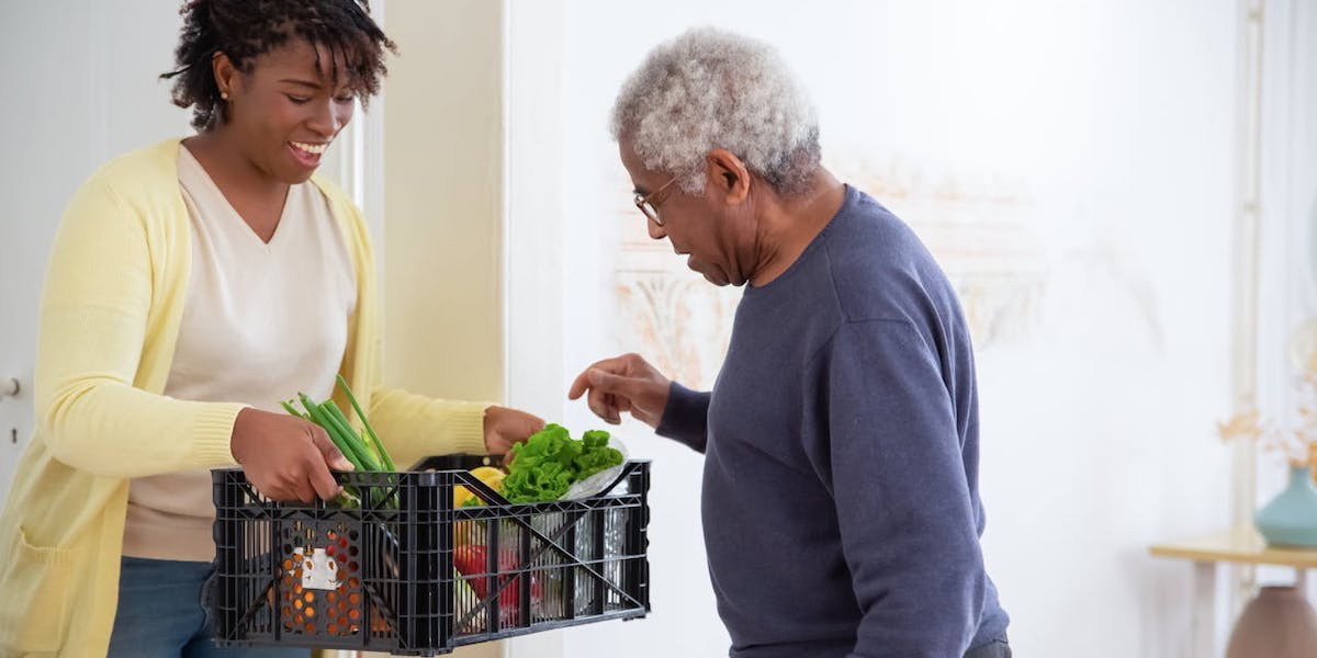 En kvinna håller fram en back med grönsaker och en äldre man tittar på dem. Du kan förbättra din hälsa så att du åldras bättre, även om du hunnit fylla 65, en växtbaserad kost är en bra start.