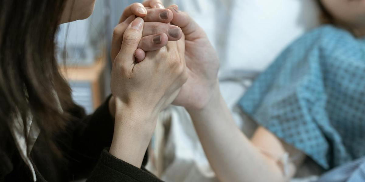 En kvinna håller handen på en kvinna som ligger i en sjukhussäng. Traumatiska upplevelser påverkar människor ända tills de dör och kan ge ökad smärta i livets slutskede.