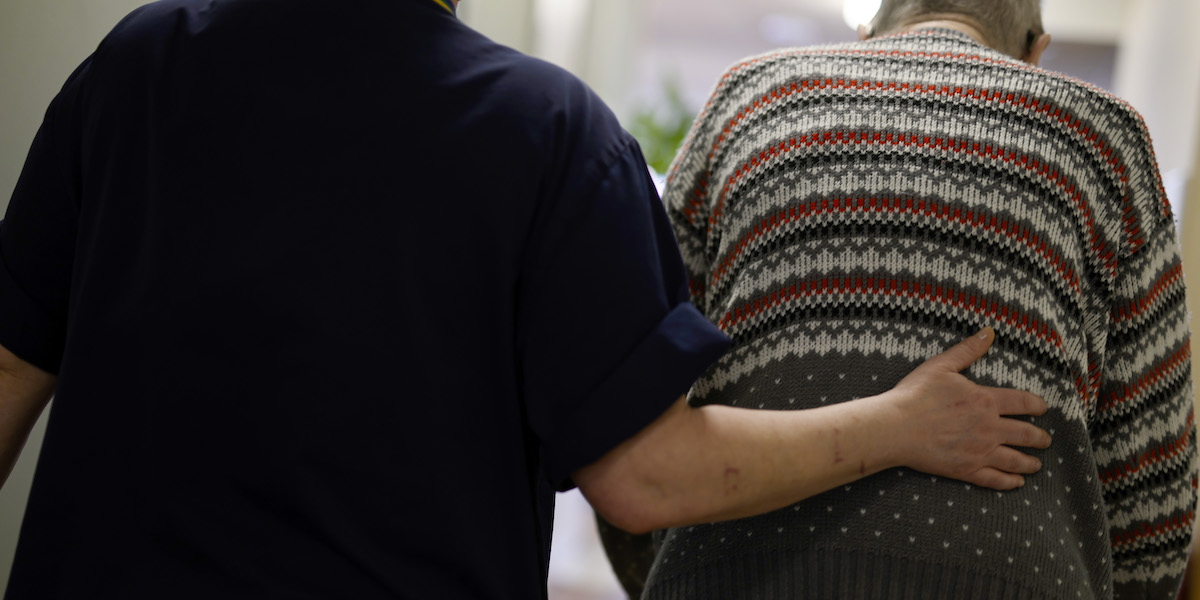 Ett vårdbiträde håller handen på ryggen på en person som bor på ett vårdboende för äldre. En utvärdering gjord av Socialstyrelsen visar att de flesta måste vänta längre än vårdgarantins 90 dagar på att få en demensdiagnos.