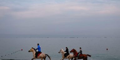Ryttare galopperar längs en strand medan oljetankrar och lastfartyg står i kö vid Hormuzsundet. 11 mars 2026.