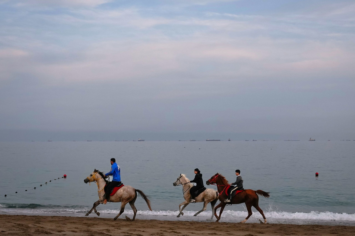 Ryttare galopperar längs en strand medan oljetankrar och lastfartyg står i kö vid Hormuzsundet. 11 mars 2026.